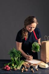 A woman in a dark apron stands by a table with fresh vegetables, herbs, nuts, and a paper bag, representing healthy eating, vegetarian cooking, and eco-friendly lifestyle.