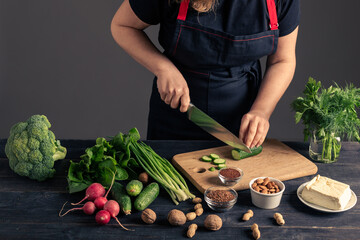 A woman in a black apron slicing fresh cucumber on a wooden cutting board. This image represents healthy eating, vegetarian lifestyle, and food preparation with organic ingredients.