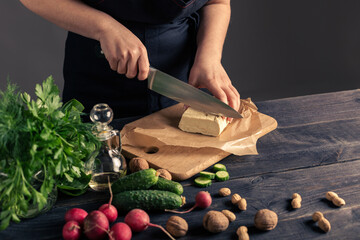 A woman in a black apron slicing tofu on a wooden cutting board. This image represents healthy eating, plant-based cooking, vegetarian lifestyle, and food preparation with organic ingredients.