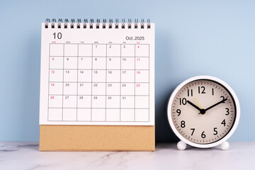 October 2025 desk calendar and analog clock on marble table with blue background, time management...