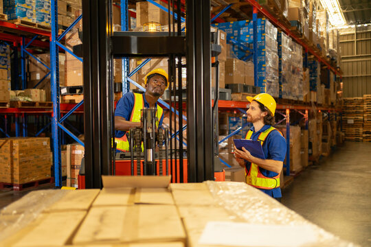 a logistics supervisor in safety gear holds documents and assesses incoming shipments against packed boxes and forklift activity in a high-bay warehouse