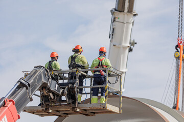 Construction workers in safety gear on a lift platform at a wind turbine installation site
