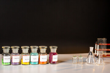 Six glass jars with multi-colored chemical compounds in powder form, labeled with chemical formulas...