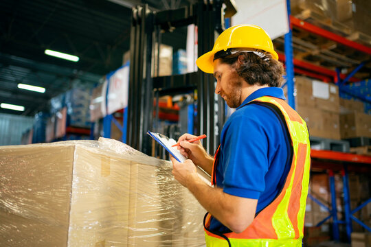 warehouse worker checking inventory list on clipboard next to pallet of boxes with forklift operator behind in a busy storage facility