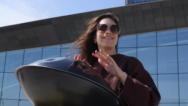 young caucasian woman playing black handpan with hands, ethnic metal drum instrument, sits on stone staircase, scyscraper and business center in background, beautiful relaxing music for meditation
