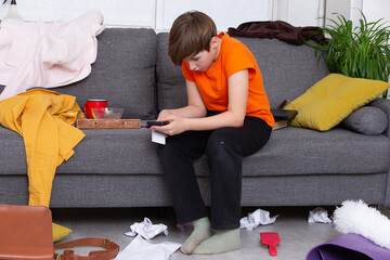 Child in orange shirt sits on messy couch using smartphone in a cluttered living room during daytime