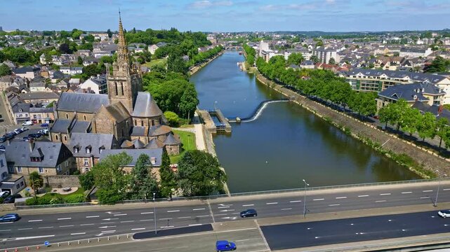 Drone flies laterally past Notre-Dame d'Avesni&egrave;res, showing Mayenne River, road, and surrounding Laval cityscape with light traffic - France