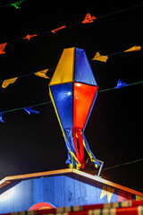 Large colorful balloon lantern in red, blue, and yellow hanging above wooden festival booth during nighttime Brazilian festa junina celebration, with decorative bunting flags strung overhead.