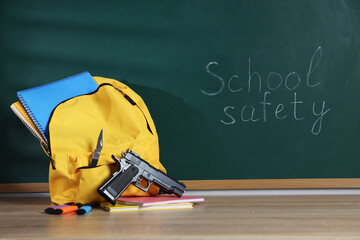 Backpack with gun, knife and stationery on wooden table near blackboard. School safety