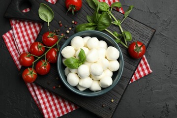 Tasty mozzarella cheese balls in bowl, basil, peppercorns and tomatoes on black table, top view