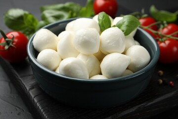 Tasty mozzarella cheese balls in bowl, basil, peppercorns and tomatoes on black table, closeup