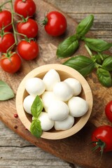 Tasty mozzarella cheese balls in bowl, basil, peppercorns and tomatoes on wooden table, top view