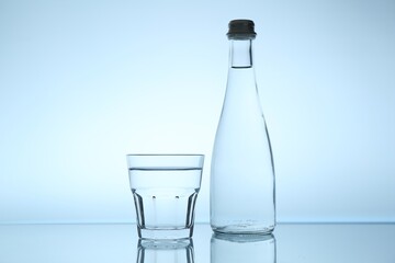 Bottle and glass of water on mirror table against light blue gradient background