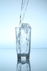Pouring water into glass at mirror table against light blue gradient background, closeup