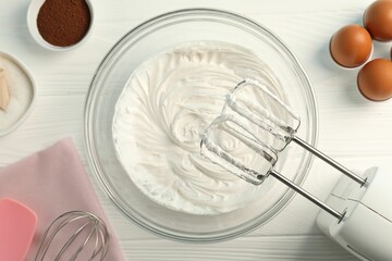 Bowl with whipped cream, hand mixer and ingredients on white wooden table, flat lay