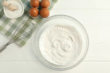 Bowl with whipped cream, whisk and ingredients on white wooden table, flat lay