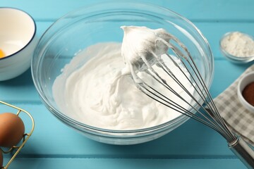 Bowl with whipped cream, whisk and ingredients on light blue wooden table, closeup