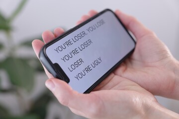 Cyber bullying. Woman holding smartphone with insults on blurred background, closeup