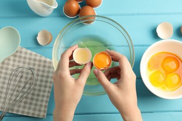 Woman separating egg yolk from white over glass bowl at light blue wooden table, top view