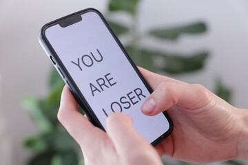 Cyber bullying. Woman holding smartphone with text You Are Loser on blurred background, closeup