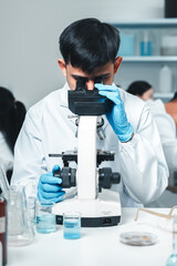 Focused Asian male scientist using microscope in modern laboratory with DNA structure chart in...