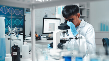 Focused Asian male scientist using microscope in modern laboratory with DNA structure chart in background. Concept of biotechnology, genetics research, molecular biology and advanced healthcare.