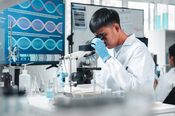 Focused Asian male scientist using microscope in modern laboratory with DNA structure chart in...