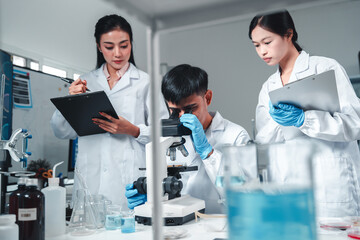 Focused Asian male scientist using microscope in modern laboratory with DNA structure chart in...