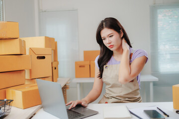 Young asian woman selling online using laptop computer and suffering from neck pain after long working hours in warehouse, surrounded by cardboard boxes