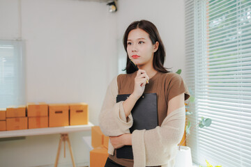 Young beautiful asian businesswoman standing holding clipboard and pen thinking about business strategy in startup office full of boxes with online orders ready for shipping