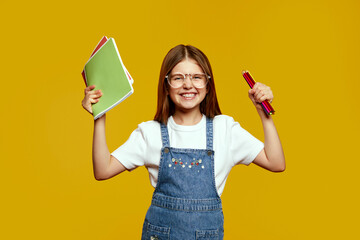 Happy kid dressed in casual overalls, enthusiastically holding notebooks and pencils, smiling...
