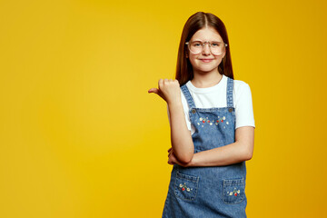 Happy schoolgirl wearing glasses and a denim dress smiles brightly, pointing confidently with her...