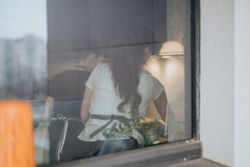 Two people, captured from outside a window, collaborating in a kitchen to prepare a meal, creating a welcoming and harmonious scene in an intimate home setting.