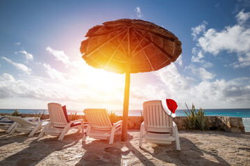 Santa Claus hat on sunbed on caribbean beach with grass umbrellas and chaise-longues. Christmas celebration
