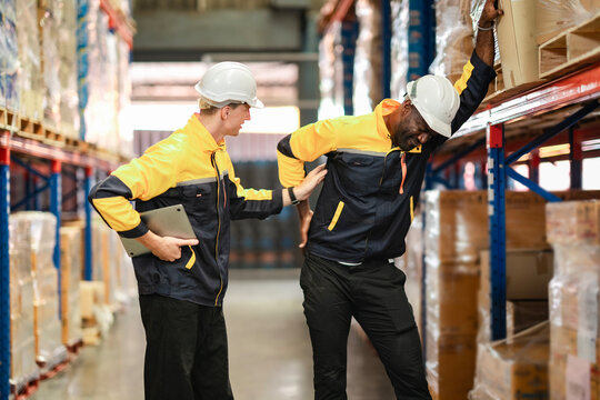 A Black man holds his lower back in pain after lifting a box, while a White coworker looks at him with concern. The image shows workplace injury risk and empathy between teammates...