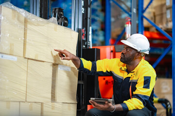 Industrial worker inspects packages in warehouse, kneeling near forklift. Using tablet for cargo...