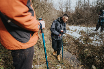 Outdoor enthusiasts hike through a cold, snowy landscape, traversing a small creek surrounded by...