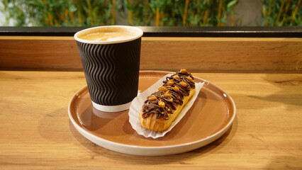 Creamy foam on cappuccino cup with french chocolate eclair topped with nuts on a table in confectionery store
