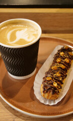 Classic Choco Eclair Sprinkled with Nuts and Coffee Latte in Paper Cup on a Clay Plate on a Wood Table