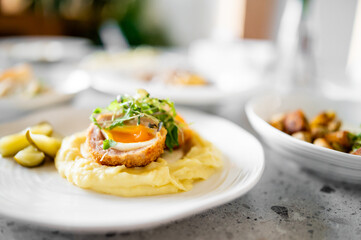 A gourmet dish featuring a breaded egg with a runny yolk on creamy mashed potatoes, garnished with fresh greens. Pickles on the side. Close-up shot with a blurred background