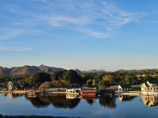 Obraz premium A peaceful riverside scene shows buildings and trees reflected in calm water, with mountains in the background and a clear blue sky above.