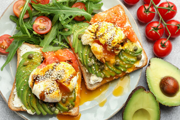 Delicious sandwiches with avocado, poached egg, salmon, tomatoes and arugula on gray table, flat lay