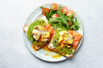 Delicious sandwiches with avocado, poached egg, salmon, tomatoes and arugula on gray table, top view