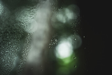 Close-up of water droplets on a fogged glass surface with soft green bokeh lighting in the background. The abstract texture and moisture pattern evoke calmness and a serene natural ambiance.