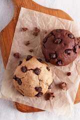 two chocolate chip cookies on parchment paper, light and dark dough, wooden board, top view