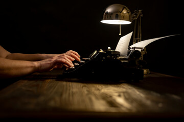 Hands typing on a vintage typewriter at night under a warm desk lamp in a cozy room creating an...