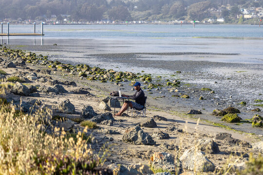 A man with a laptop working on a beach or bay at low tide.  Full-time RV nomad lifestyle, working in shorts from his home office.