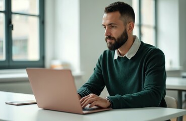 Bearded man works on laptop at desk in bright office. Business pro looks at computer screen. Male in green sweater. Remote work, online communication, modern workplace, corporate environment.