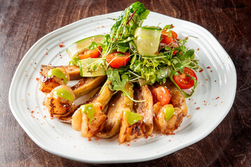 Grilled shrimp and eggplant with a fresh salad of arugula, cherry tomatoes, and cucumber ribbons, garnished with sesame seeds and green sauce, served on a wooden table.