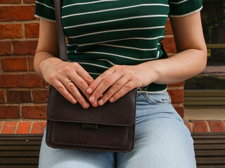 A girl in blue jeans sits on a bench, a handbag on her lap, handles on it